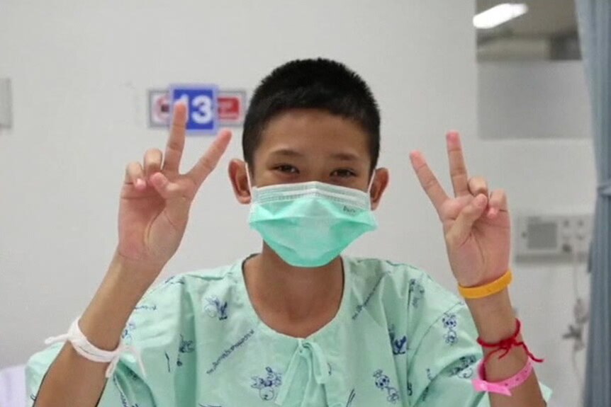 Rescued Thai boy gives two peace signs while in hospital gown and mask