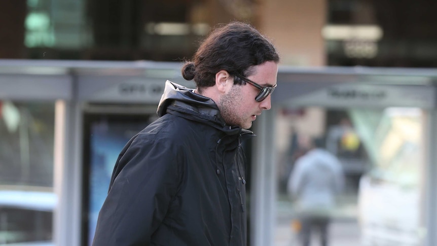 Brent Moresco, wearing a black hooded jacket and sunglasses, walks in a city street.