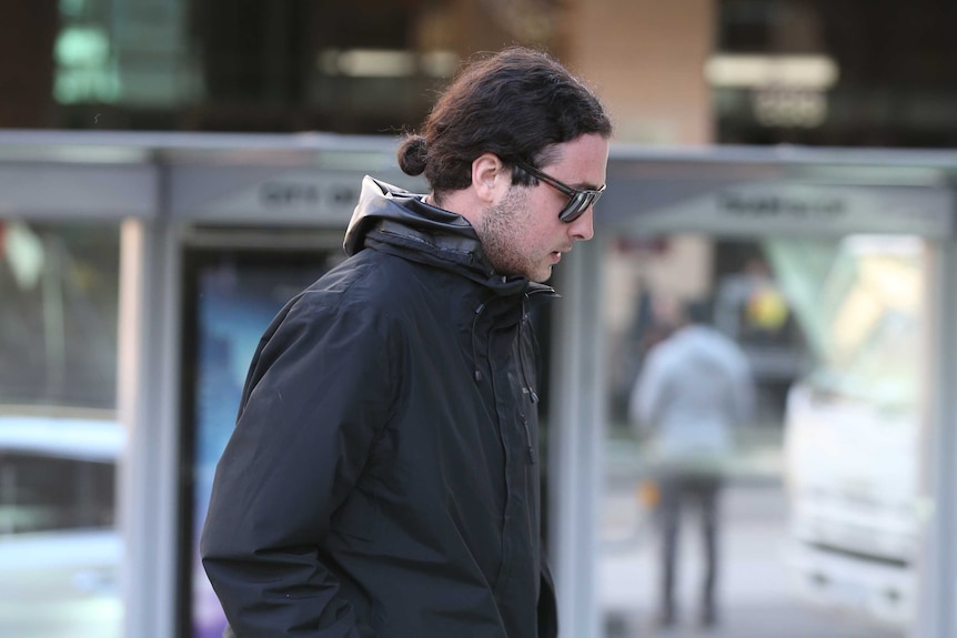 Brent Moresco, wearing a black hooded jacket and sunglasses, walks in a city street.