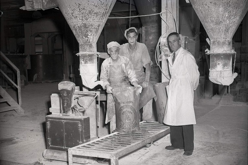 Dusty workers inside bagging plant with a bag of titanium oxide.