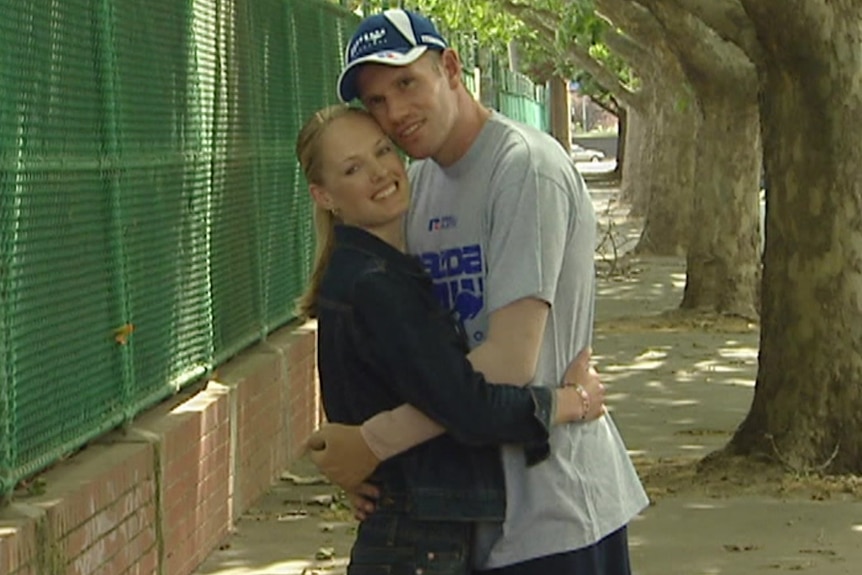 A woman in a black top holds a man in North Melbourne cap and shirt on a sunny day. There is gauze on his arms.