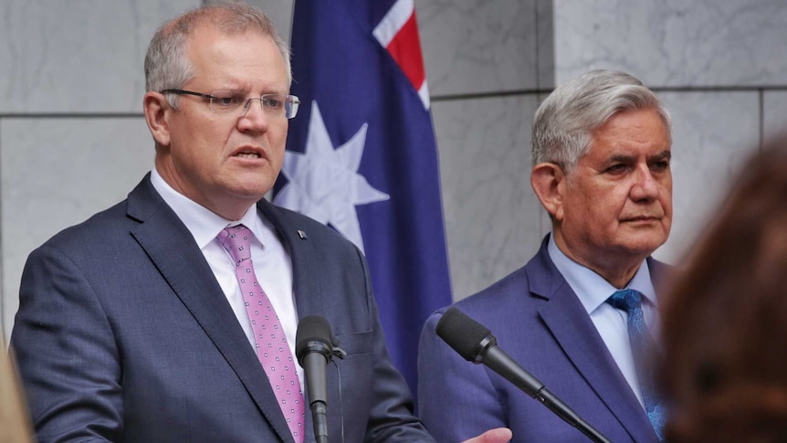 Two men speak at a press conference in front of a flag