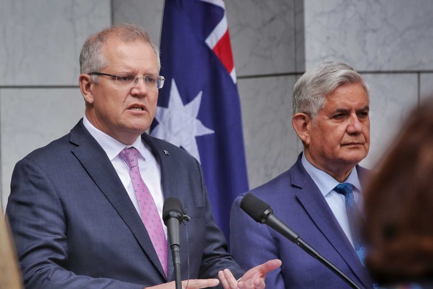 Two men speak at a press conference in front of a flag