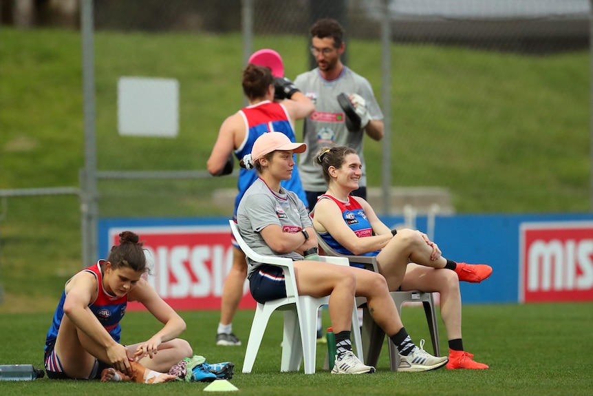 Ellie Blackburn and Kirsten McLeod sit in chairs beside the boundary line as their teammates train