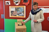 A woman in a light grey jacket and a blue scarf stands beside a voting box .