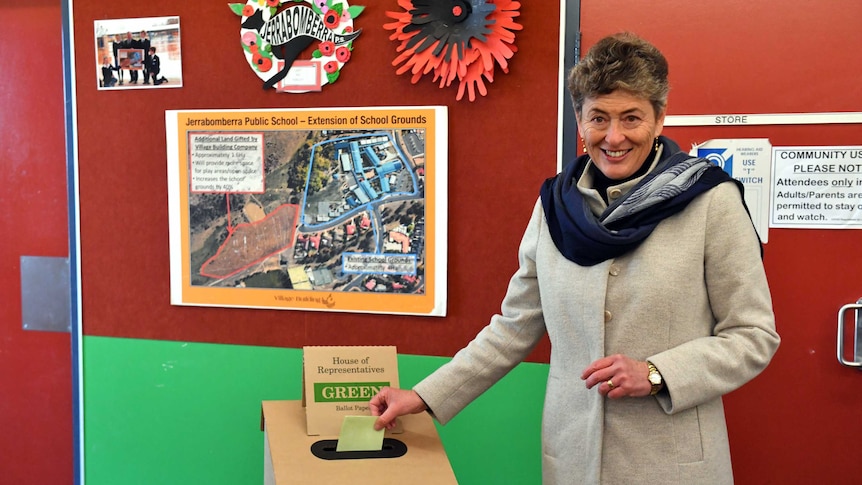 A woman in a light grey jacket and a blue scarf stands beside a voting box .