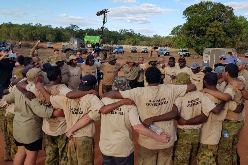Group of men in Kapani Warrior t-shirts and khakis stand in a circle with their arms around each other's shoulders
