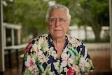 An elderly man with a button-up shirt with flowers stares at the camera.