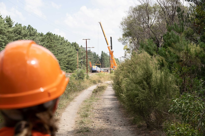 A crane picks up part of a derailed train, behind a person wearing an orange hard hat.