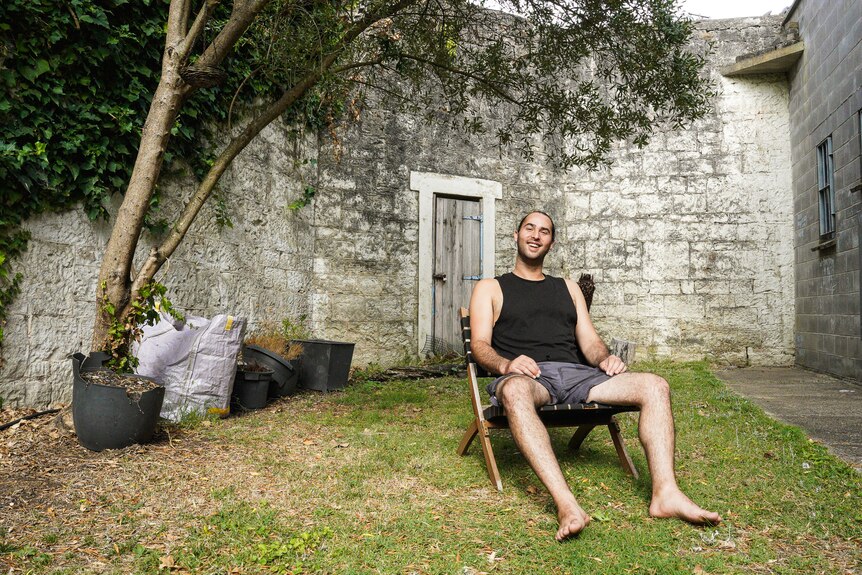 A man wearing a black singlet and board shorts sits on a fold out chair in a grassy courtyard.