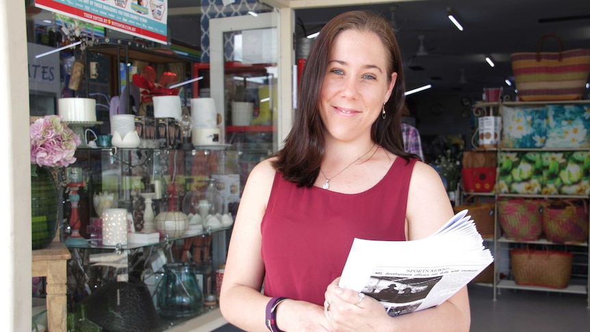 A woman smiling in front of a newsagency, holding newspaper.