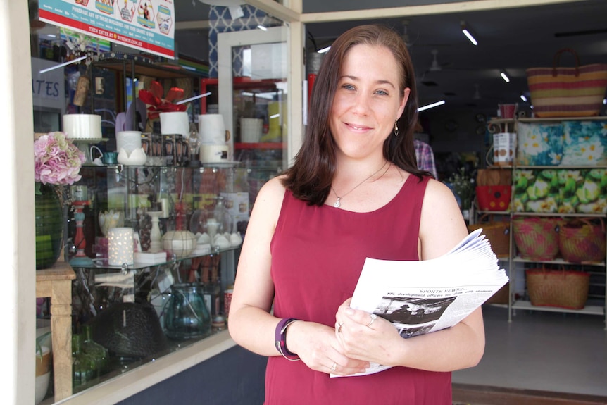 A woman smiling in front of a newsagency, holding newspaper.
