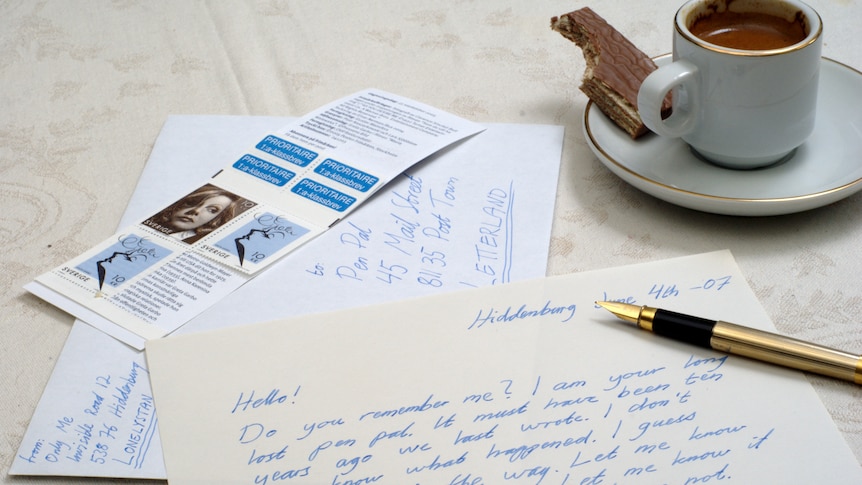 A partially-written pen-pal letter with a gold pen on top, stamps and an envelope, with an espresso and chocolate biscuit above.
