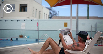 Man in bathers on banana lounge reads newspaper under and umbrella next to a public swimming pool, visible in background.