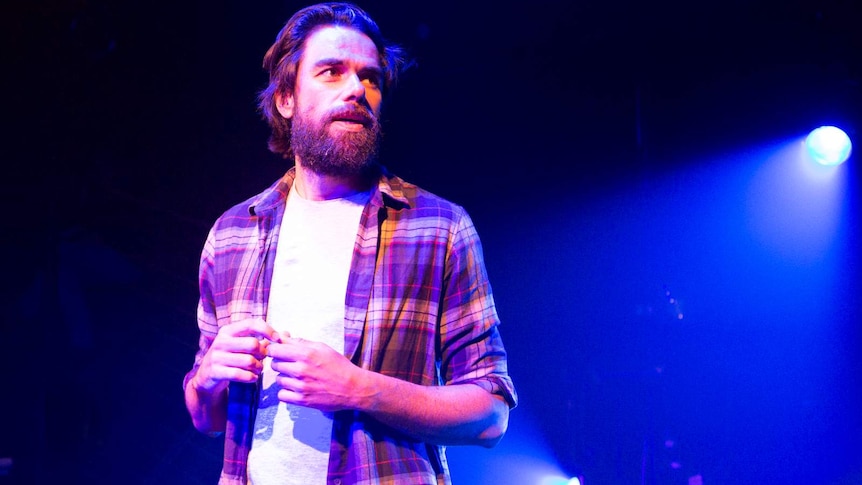 Actor with beard and moustache wearing flannel shirt over a white t-shirt stands on dark stage, spot-lit from behind.