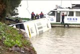Four people in life jackets try to retrieve the wrecked bus, resting in a shallow river