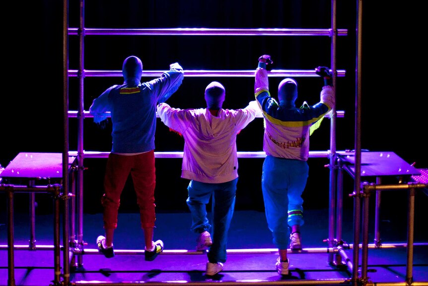 Three people climb a small scaffolding wall on a stage.