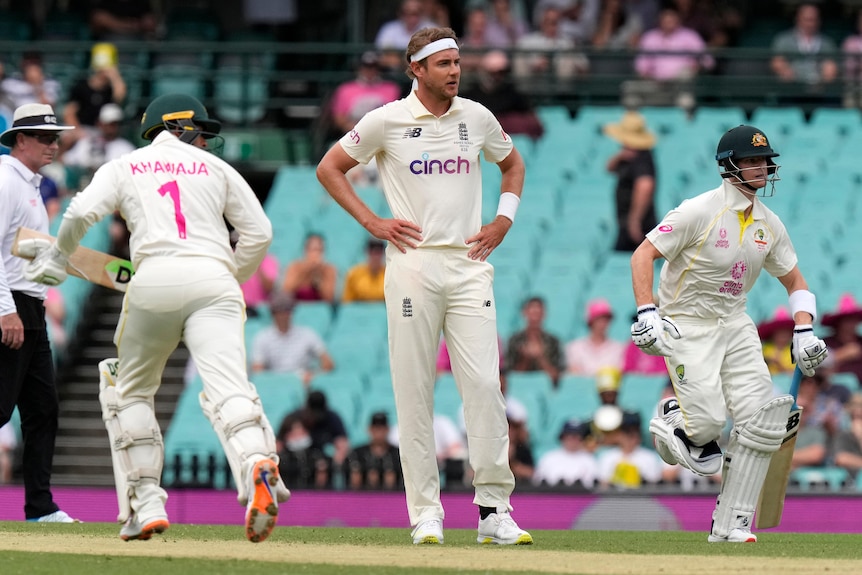 England bowler Stuart Broad stands with hands on hips as Australia batters Usman Khawaja and Steve Smith run between wickets.