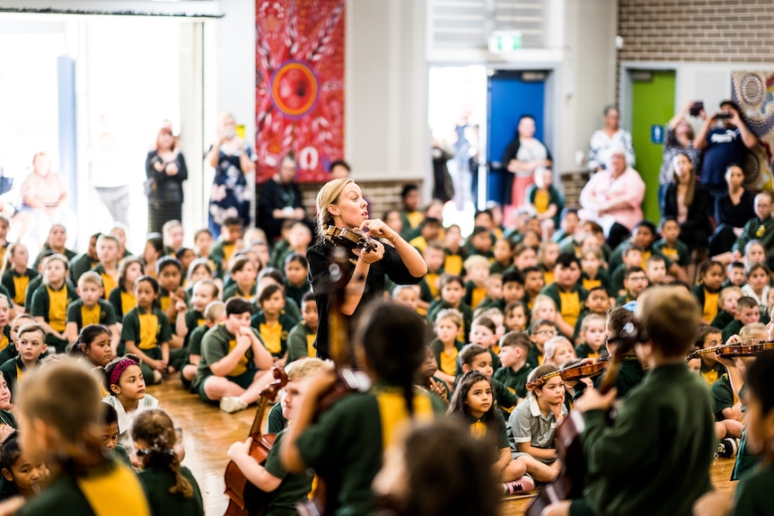 A white woman playing a violin stands in a roomful of primary school students, leading an ensemble of string players.