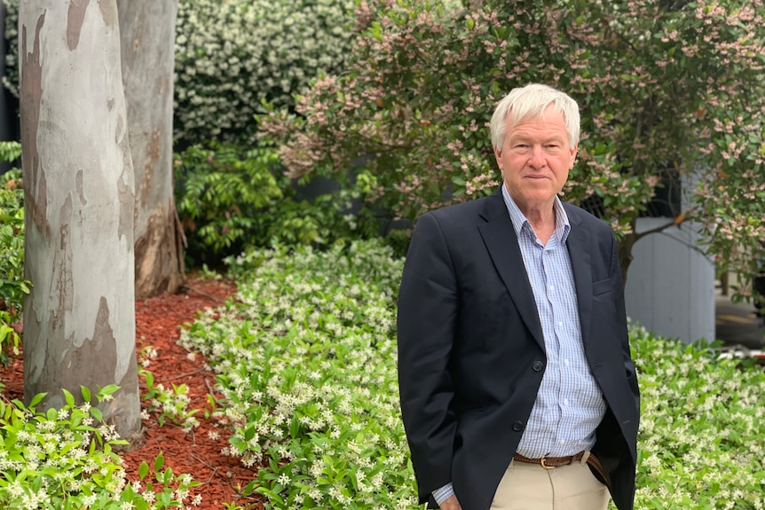 A man with white hair, a blue shirt, blue blazer and brown pants standing in front of bushes and a tree trunk.