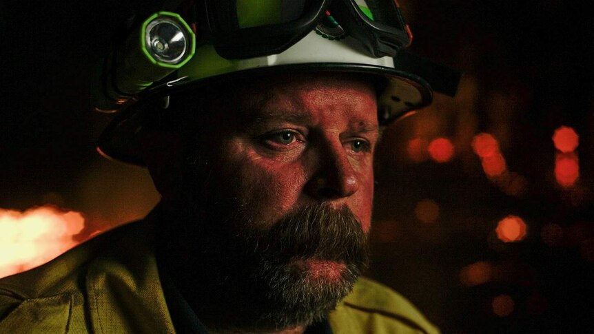 A close-up of firefighter seen wearing yellow jacket and white helmet with torch and goggles. He stares into space.