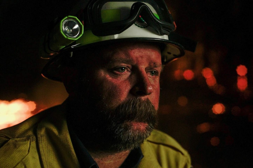 A close-up of firefighter seen wearing yellow jacket and white helmet with torch and goggles. He stares into space.