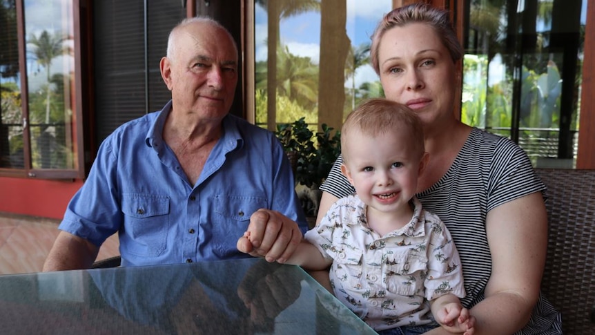 Ron Rutland sitting at a table on his deck with his daughter Amanda Rutland and smiling 22-month-old grandson Naish Dodson.