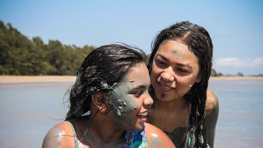 Two young Gunnamulla girls, painted with grey clay, smile at each other on the beach.