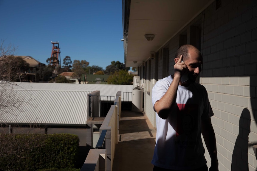 Josh ?? talks on phone, his shadow cast by the sun against the wall and Kalgoorlie's ?? visible against the blue sky behind him.