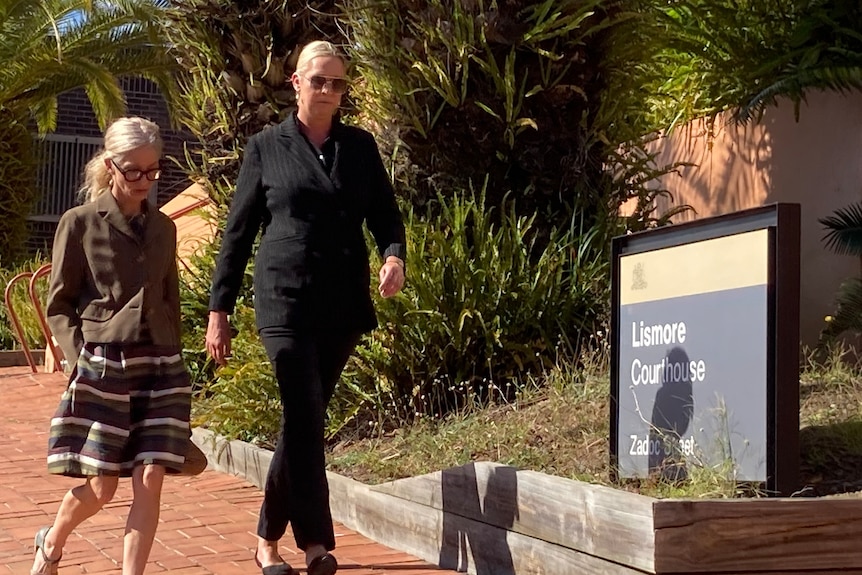 A blonde woman in a back outfit walks past a Lismore Courthouse sign