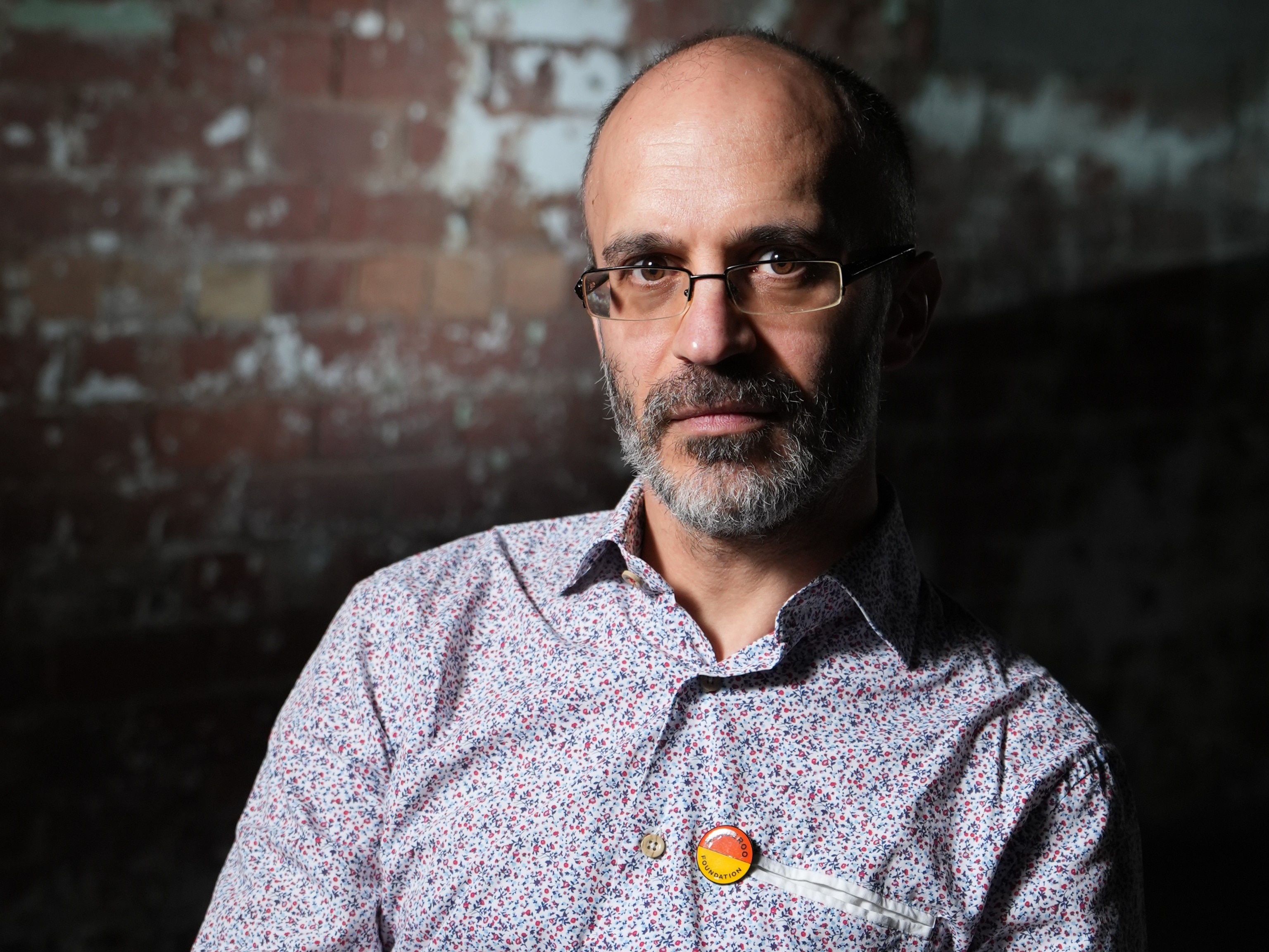 A man with frameless glasses, greying beard and paisley business shirt in front of a brick wall.