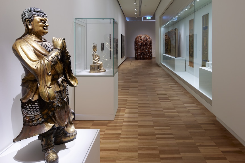Side-on photo of a gold Chinese Buddha statue on a white box, with other gallery exhibits just visible in background.