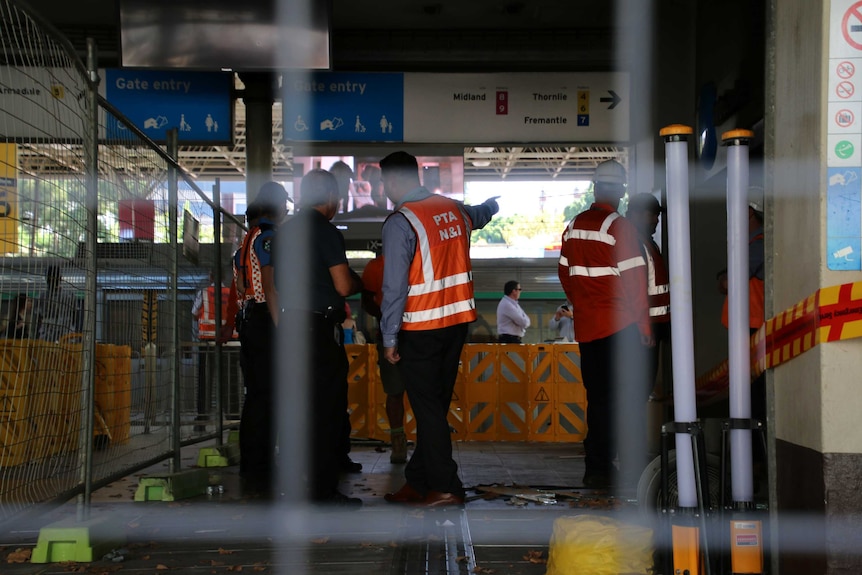 A group of people in hi-vis clothing stand behind a wire fence at Perth Train Station examining damage from a fire.