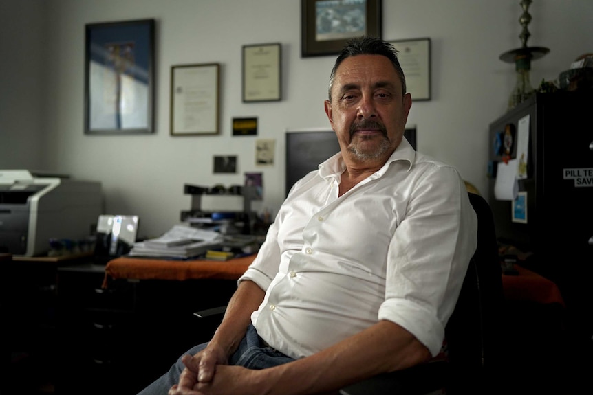 Man wearing white shirt sits in office with framed documents on wall behind him