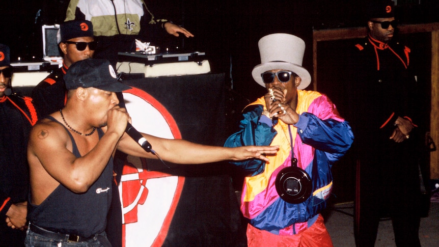 Chuck D wears tank top, Flavor Flav wears colourful tracksuit and white top hat. Both rap on stage as DJ plays behind them.