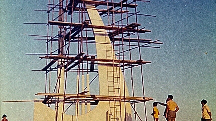 Grainy historical image of scaffolding around a white frame of a sail boat with people watching on.