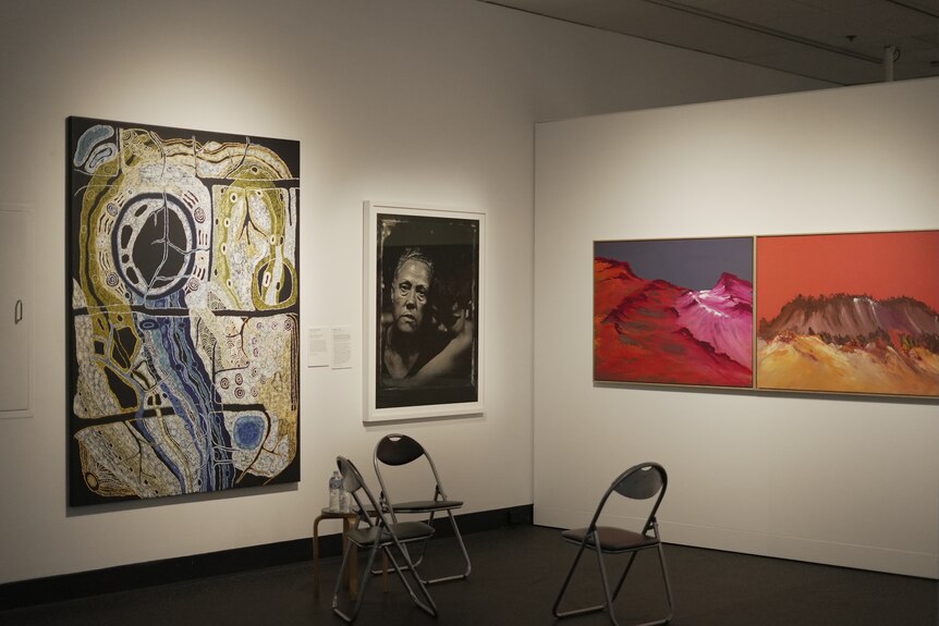 Three large artworks on two walls in a gallery. Three black chairs sit in front of them