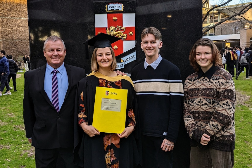 A young woman stands in university graduating robe and hat, with her parents and brother around her.