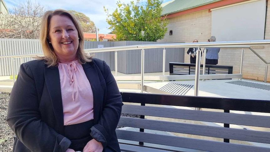 A woman in a pink top and black blazer sits on a bench outside 