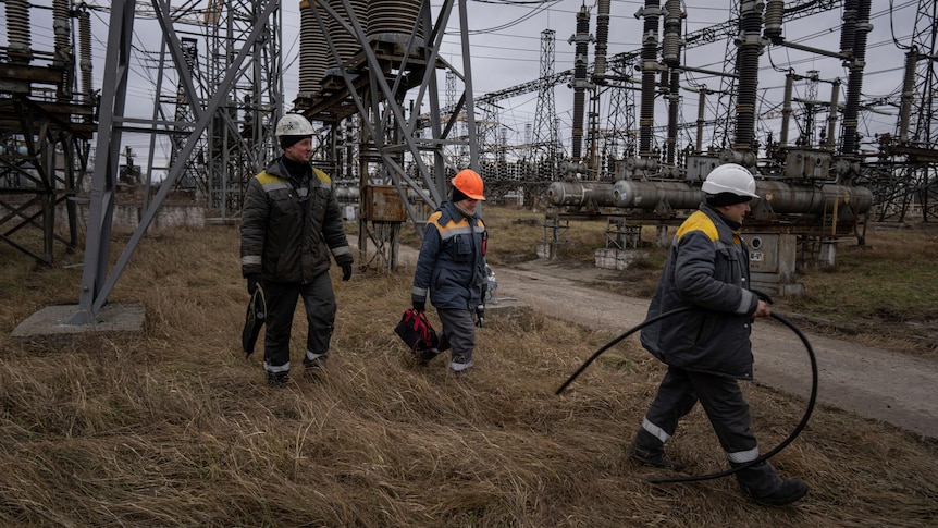 three power workers in hard hats and protective gear walk through a power plant outside in Ukraine