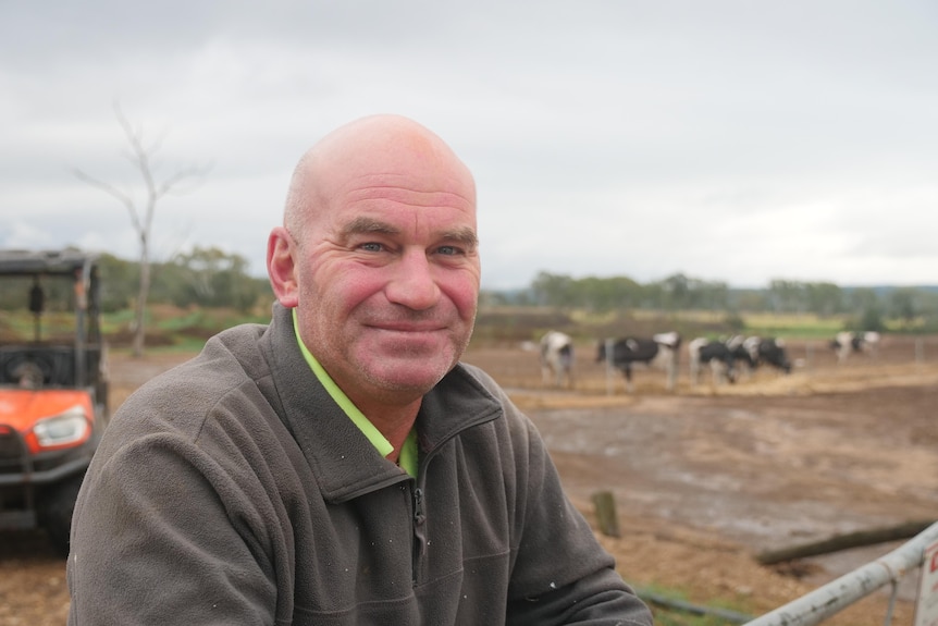 Nanango dairy farmer Illya Childs smiles with a sodden paddock and cattle in the backgound.