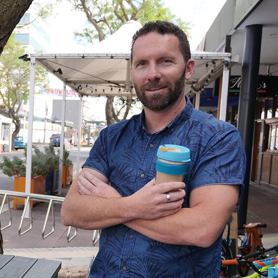 A man with short dark hair and a beard, wearing a blue shirt and holding a coffee, standing in a town street.