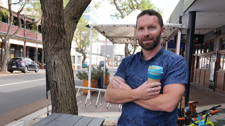 A man with short dark hair and a beard, wearing a blue shirt and holding a coffee, standing in a town street.