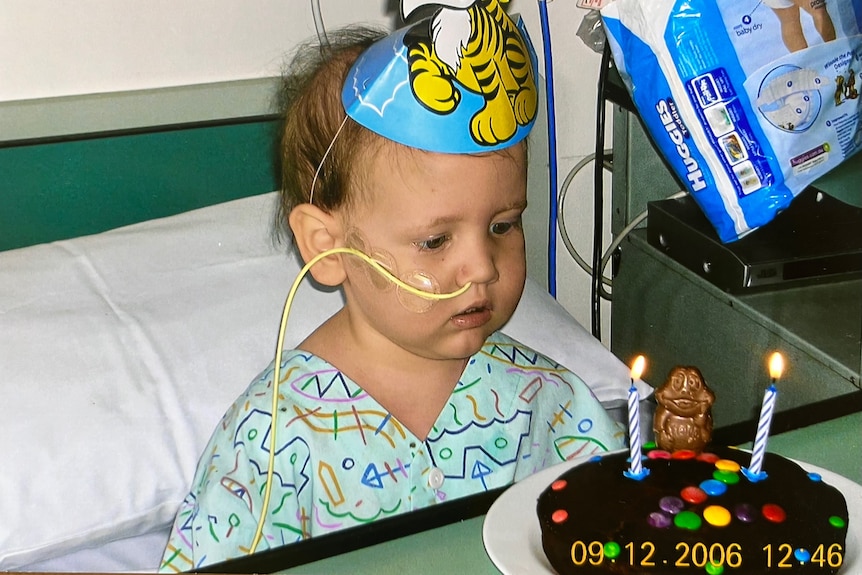 A young boy wears a party hat and blows out the candles on his birthday cake from a hospital bed