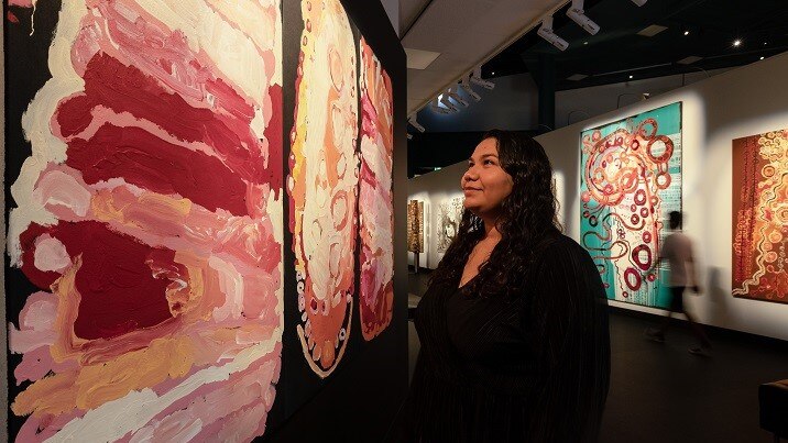 A woman standing in front of a painting, admiring it. The painting uses pink and white hues on a black background.