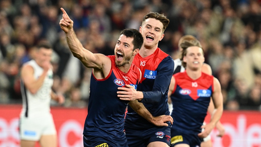 A Melbourne AFL player points to fans in celebration as his teammates congratulate him after a goal.