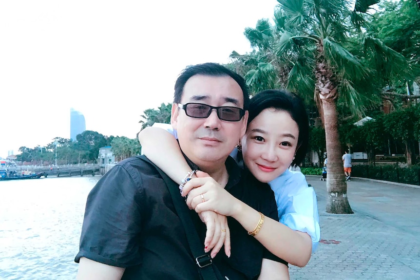 A man in a black shirt sits beside water while a woman in a blue shirt puts her arms around his neck.