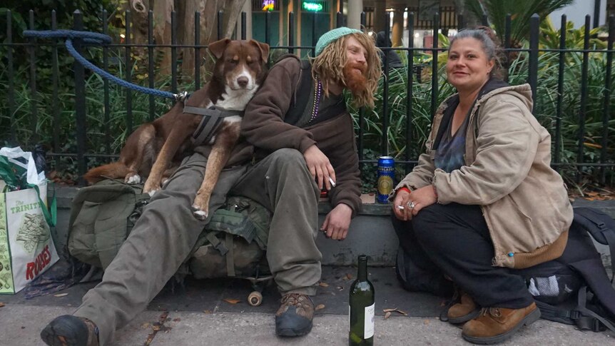 Two homeless people with a dog share a drink and a smoke