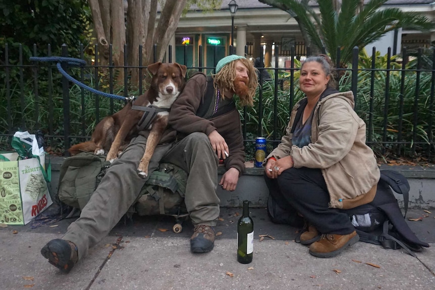 Two homeless people with a dog share a drink and a smoke