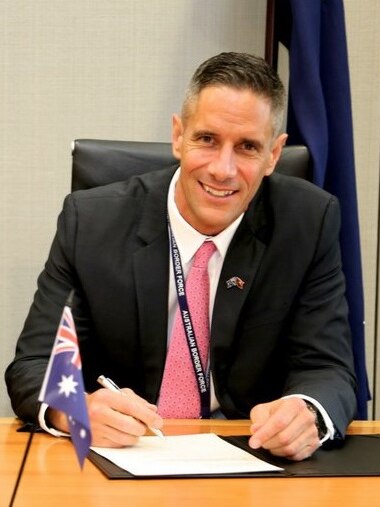 Roman Quaedvlieg smiles at the camera while signing a piece of paper. There is a small Australian flag on a stick in front.